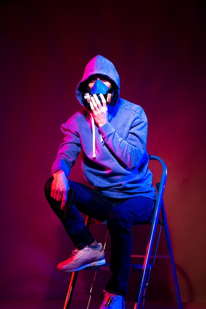 Portrait of an anonymous man, hacker wearing neon mask over dark background. Selective focus of bi-racial cyberpunk player holding guns near neon lighting