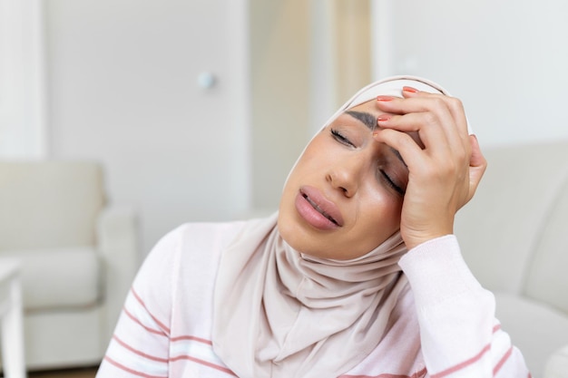 Portrait of an arabic muslim woman sitting on a sofa at home\
with a headache feeling pain and with an expression of being unwell\
upset woman lying on couch feeling strong headache migraine