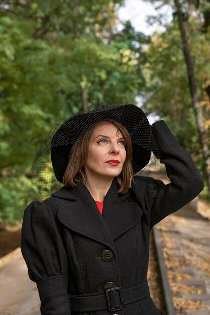 Portrait of aristocratic woman in black hat with veil looking up in autumn park Middleaged woman in vintage retro style