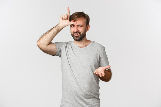 Portrait of arrogant man in gray t-shirt, mocking person who lost, showing loser sign on forehead