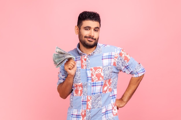 Portrait of arrogant wealthy bearded man in casual style blue shirt standing showing wad of money looking at camera with proud expression income Indoor studio shot isolated on pink background