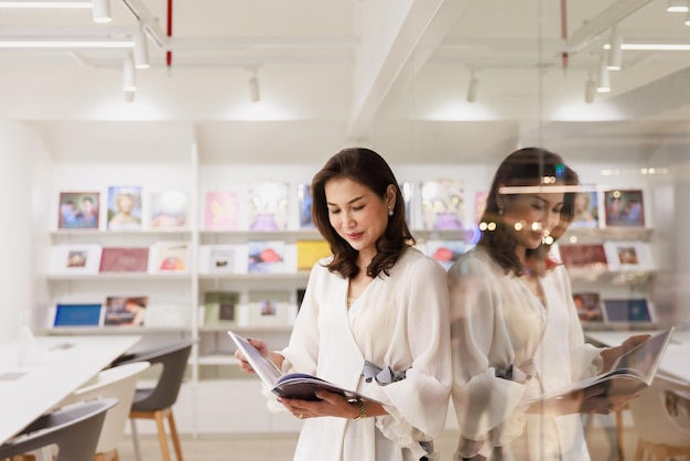 Portrait of Asain pretty cheerful female standing and reading book in library and lean on glass wall with happy focus and concentrate with reflection in clear mirror