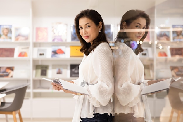 Portrait of Asain pretty cheerful female standing and reading book in library and leaning on glass wall with happy focus and concentrate with reflection in clear mirror looking at camera