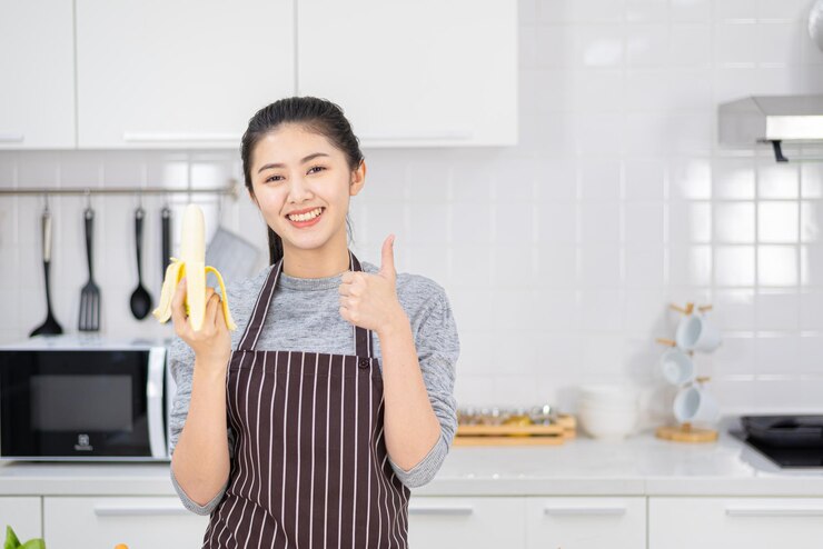 Photo アジアの美しい女性と主婦の肖像画は笑顔で白いキッチンでバナナを保持しています