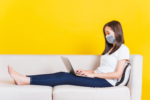 Portrait Asian of beautiful young woman sitting on sofa wearing medical face mask protective she work from home with laptop computer during Coronavirus studio shot isolated on yellow background