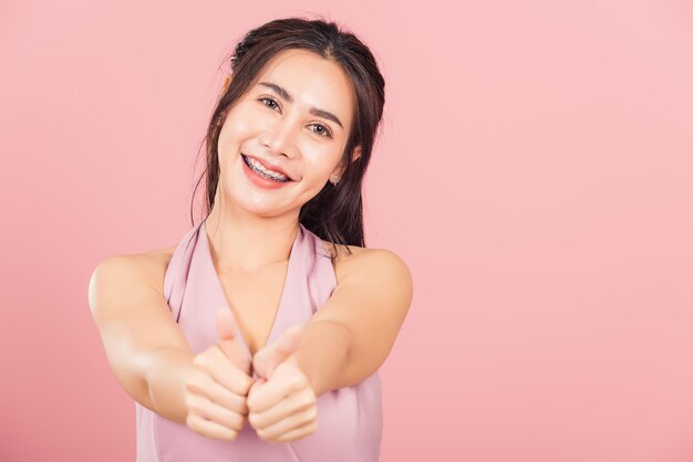 Portrait Asian beautiful young woman smile she standing made finger thumbs up, good sign to agree studio shot isolated on pink background, Thai female successful like finger gesture with copy space