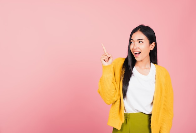 Portrait Asian beautiful young woman smiling standing pointing finger out on pink background, Thai happy face excited female point into empty looking to side away with copy space for text