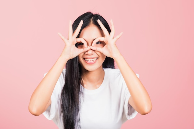 Portrait Asian beautiful young woman standing, She made finger OK symbol sign to agree near eyes looking at camera, studio shot isolated on pink background, Thai female agree hand sign copy space