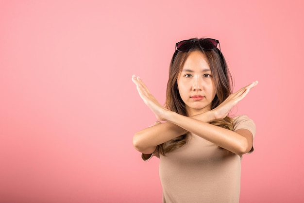 Portrait Asian beautiful young woman unhappy or confident standing holding two cross arms say no X stop sign, studio shot isolated pink background, Thai female pose reject gesture with copy space