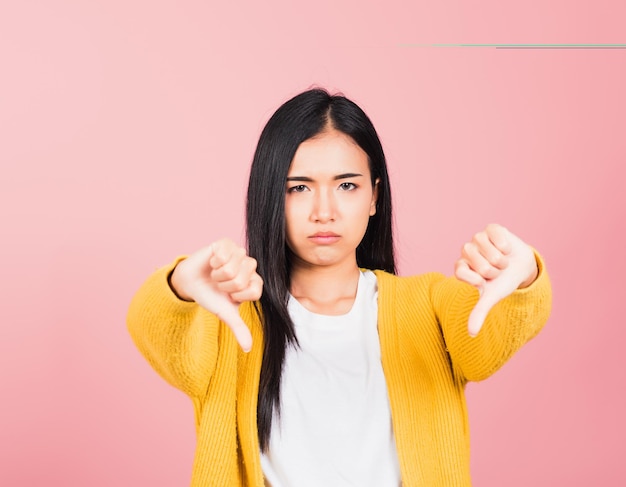 Portrait Asian beautiful young woman unhappy, negative gesture showing finger thumbs down or dislike sign, studio shot isolated on pink background, Thai female rejection unlike with copy space