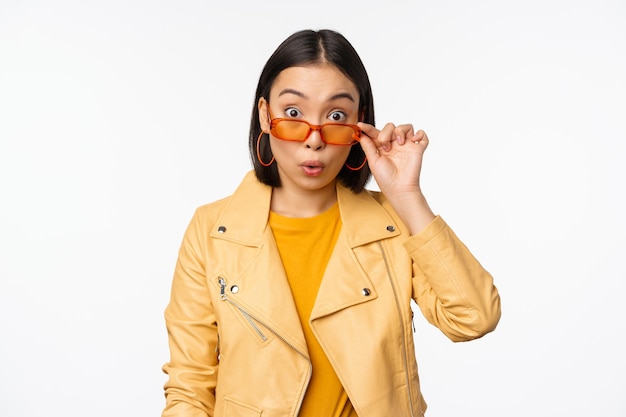 Portrait of asian brunette woman in stylish sunglasses looks surprised and impressed at camera checking out big news wow face expression standing over white background