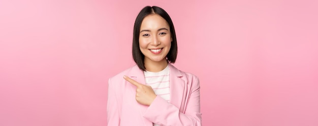 Portrait of asian business woman saleswoman in suit pointing finger left showing banner advertisement smiling and looking professional pink background