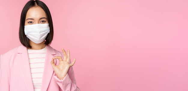 Портрет азиатской бизнесвумен в медицинской маске, показывающей знак "хорошо" в костюме, правила работы во время