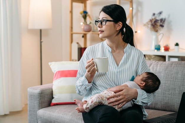 La madre asiatica della donna di affari del ritratto che tiene il suo bambino che dorme sta guardando in lontananza mentre fa una pausa caffè dal lavoro in una giornata tranquilla nel soggiorno