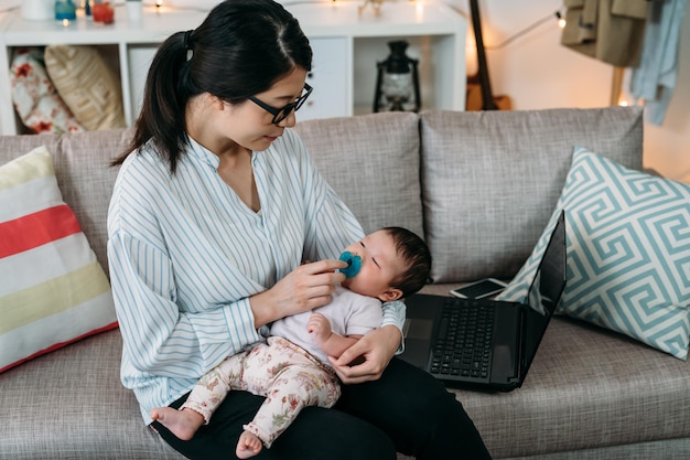 portrait asian career woman working from home is keeping hand on the pacifier while her lovely new baby is sleeping peacefully in arms on the living room sofa.