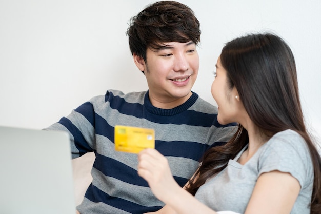 Portrait of a asian cheerful couple shopping online on internet with laptop computer while sitting at on the bed at home, man holding credit card with feeling happy for payment a gift to his wife