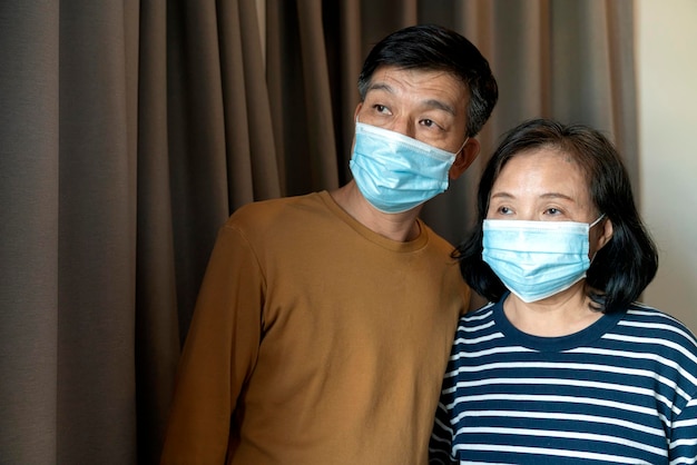 自宅で顔の医療マスクパンデミックコロナウイルス病検疫を身に着けているアジアのカップルの年配の老人男性と女性の肖像Covid19発生防止の概念