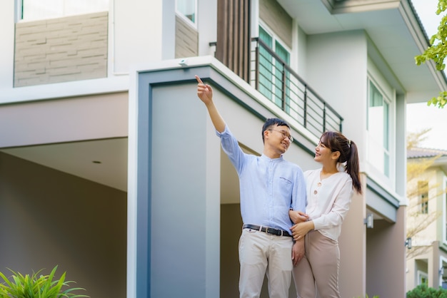 Portrait of Asian couple walking hugging and pointing together looking happy in front of their new house to start new life. Family, age, home, real estate and people concept.