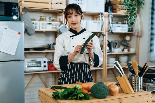 ポートレートのアジア人女性料理人が、素朴なキッチンでオンラインの料理動画を撮影しながら、新鮮な野菜の選び方のヒントを示しています。