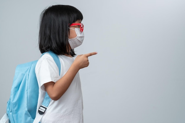 Portrait of Asian girl kid with protective face mask with school backpack and point the finger Concept of kid going back to school and new normal lifestyle