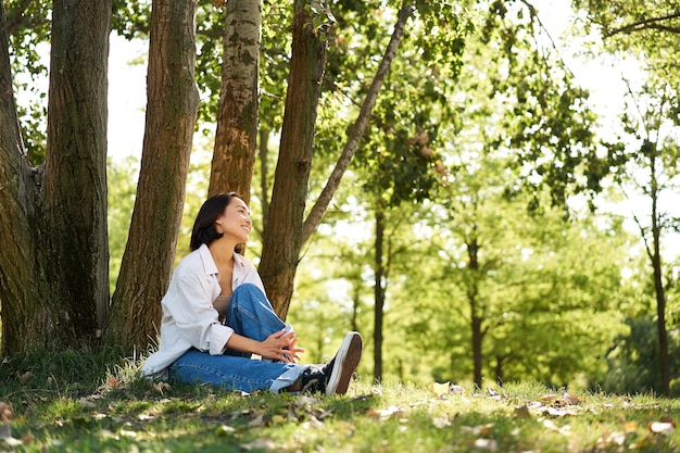 木にもたれてリラックスし、日陰の公園で休んで笑顔で楽しんでいるアジアの女の子の肖像画