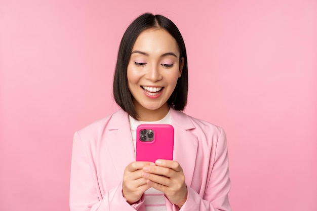 Portrait of asian girl in suit with smartphone smiling and looking happy standing over pink studio background Copy space