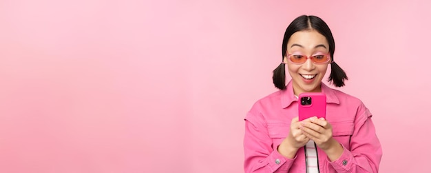 스마트폰을 사용하여 선글라스를 쓴 아시아 소녀의 초상화 분홍색 배경 위에 서 있는 앱에서 휴대전화 검색을 보고 있는 여성