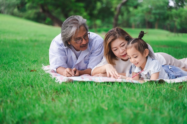 Portrait of Asian grandparents and granddaughter laying on the green glass field outdoor, family enjoying picnic together in summer day concept