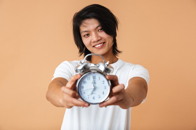 Portrait of asian guy wearing white t-shirt smiling and holding alarm clock while looking at camera isolated over beige wall