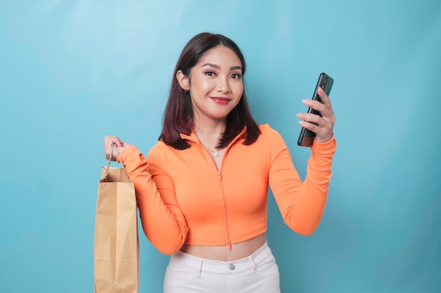 Portrait Asian happy beautiful young woman standing excited holding online shopping bag and smartphone on hand studio shot isolated on blue background