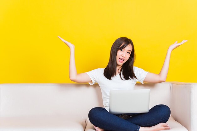 Portrait asian of happy beautiful young woman work from home she sitting on sofa using laptop computer in house living room raise hand up away studio shot isolated on yellow background