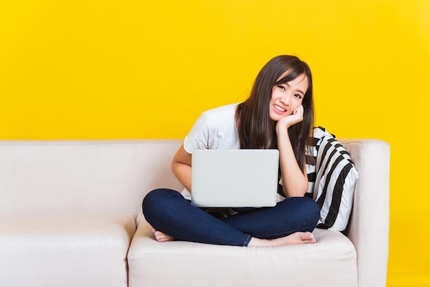 Портрет азиатки счастливой красивой молодой женщины, работающей из дома, она сидит на диване с помощью портативного компьютера в гостиной студии, снятой на желтом фоне