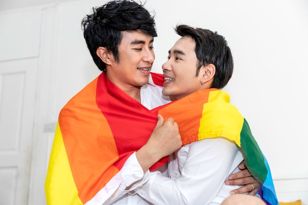 Portrait of Asian homosexual couple hug and holding hand with pride flag in bedroom