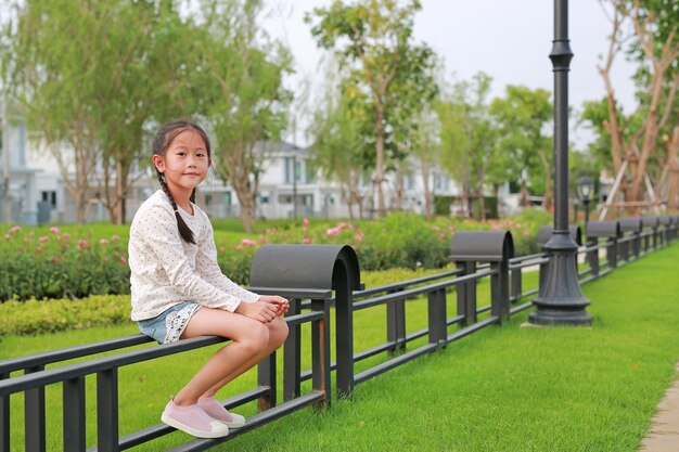 屋外の公共公園の庭のフェンスに座っているアジアの子供のポートレート