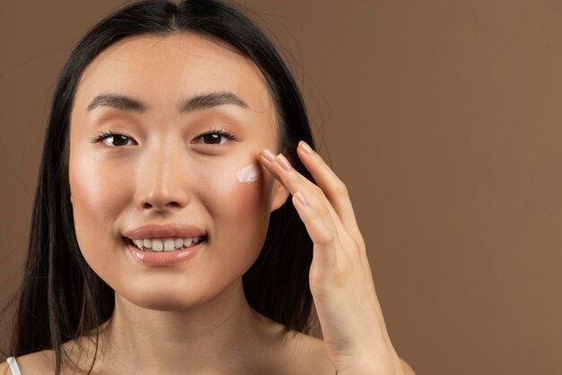 Photo portrait of asian lady applying antiaging or moisturising face cream posing over brown studio background copy space