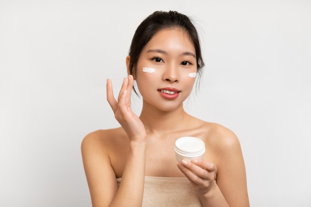 Portrait of asian lady with cream on cheek holding jar