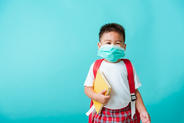 Портрет азиатских маленький ребенок мальчик детский сад носить защитную маску и школьную сумку держать книгу, прежде чем идти в школу