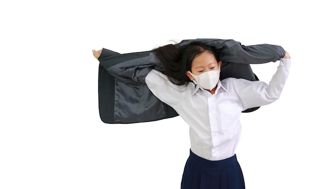 Portrait of Asian little girl child in white shirt with protective face mask raise hand up to taking off formal student suit isolated on white background. Image with Clipping path