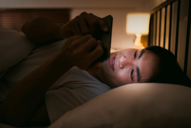 Portrait of Asian man lying in bed at night and using smartphone Young man lying in bed