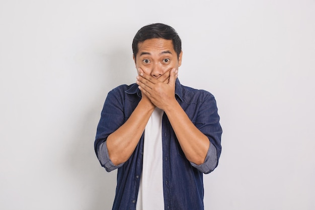 Portrait of asian man with surprised expression covering mouth with two hands on yellow background