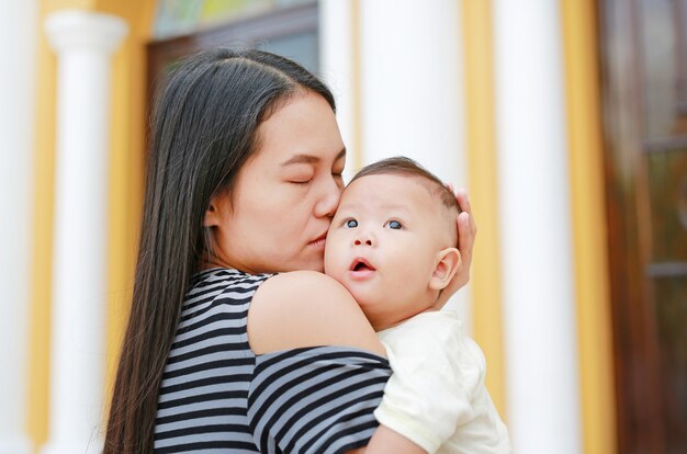 屋外の彼女の幼児の赤ちゃんを運ぶとキスをするアジアの母親の肖像画。