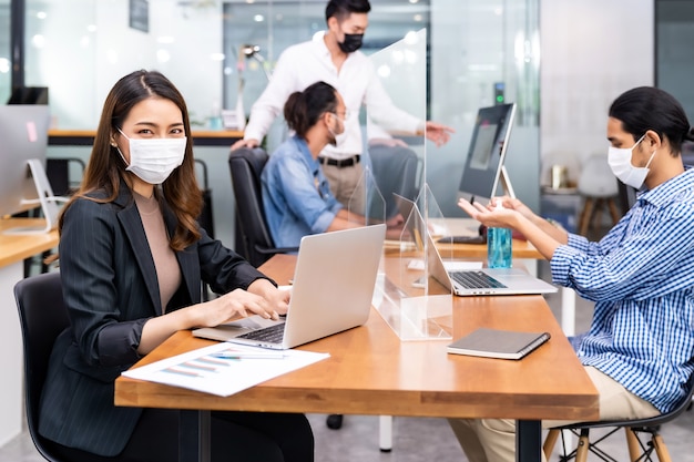 Portrait of asian office employee businesswoman wear protective face mask work in new normal office with interracial team in background as social distance practice prevent coronavirus COVID-19.