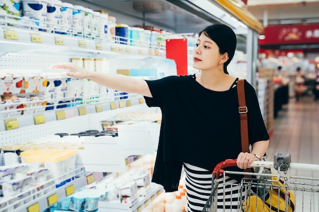 肖像画アジアの妊娠中の女の子はスーパーマーケットのミルク部門でヨーグルトを取ります。ハイパーマーケットの冷蔵庫からの製品のショッピングカートを持つスタイリッシュな母性女性。食料品店で商品を購入する