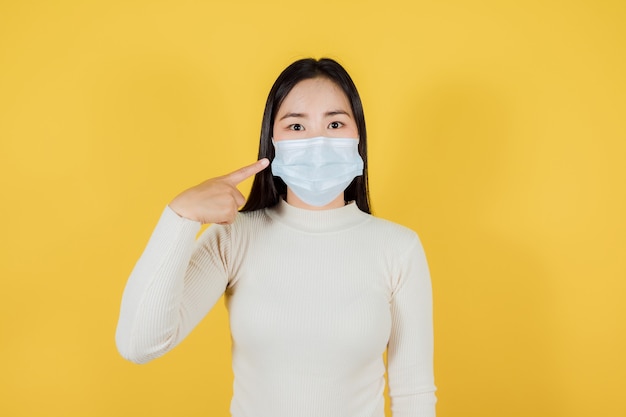 Portrait of asian teenager wearing and pointing a medical face mask to protect Covid-19 (Coronavirus) on yellow background