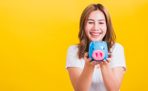 肖像画アジアのタイの美しい幸せな若い女性の多くのお金で青い貯金箱を保持し、piggybank、コピー スペースで黄色の背景に分離されたスタジオ ショットを笑顔