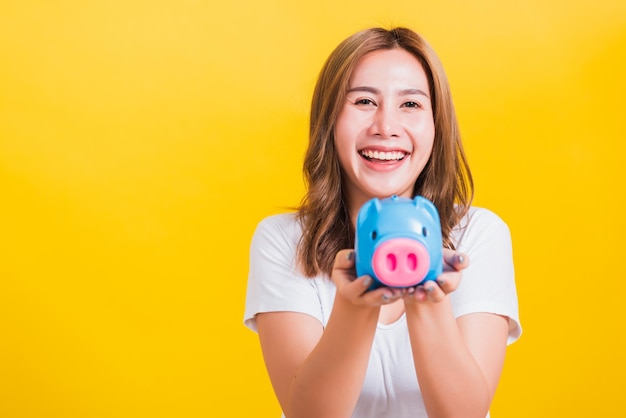 Portrait asian thai beautiful happy young woman smiling holding piggy bank with a lots money and looking to camera, studio shot isolated on yellow background, with copy space