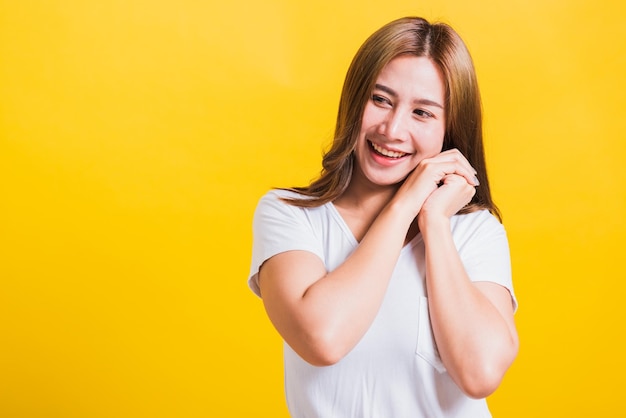 Portrait asian thai beautiful happy young woman smiling, screaming excited keeps two hands together near the face and looking to side, studio shot isolated on yellow background, with copy space