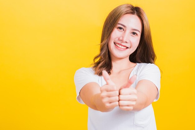 Portrait asian thai beautiful happy young woman smiling wear white t-shirt standing successful woman giving two thumbs up gesture sign in studio shot, isolated on yellow background with copy space