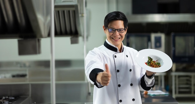 Foto ritratto di un cuoco asiatico thailandese che guarda la telecamera con il pugno in su e mostra il cibo in piatto bianco lezione di cucina classe di cucina scuola di cucina lezioni di cucina in una scuola di cucina