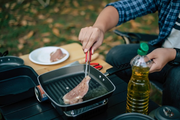 Portrait of Asian traveler man glasses pork steak frying BBQ in roasting skillet pan or pot at a campsite Outdoor cooking traveling camping lifestyle concept